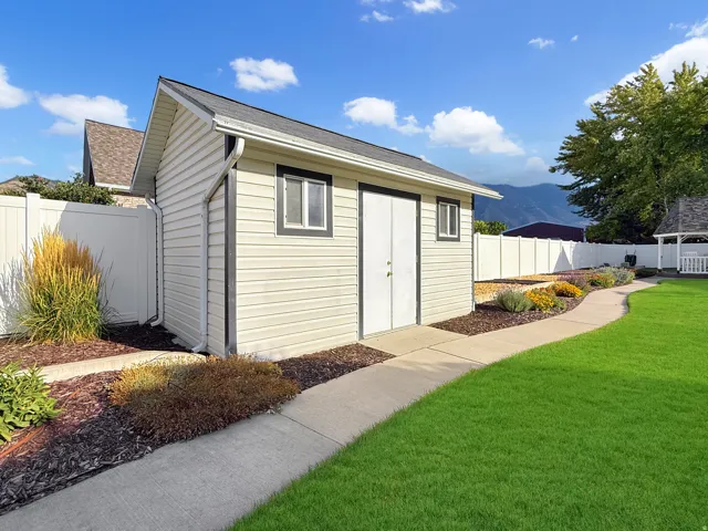 Detached storage building with dual entry doors, surrounded by a concrete pathway, manicured lawn, and planted garden beds