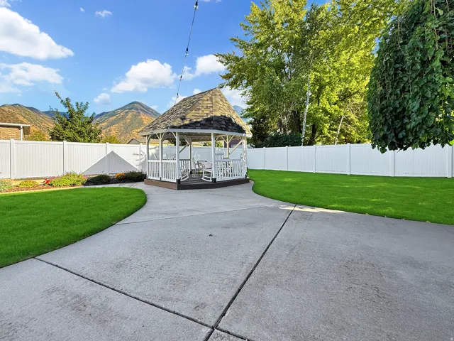 Gazebo structure with a shingled roof and white railing, surrounded by a concrete patio and a manicured lawn