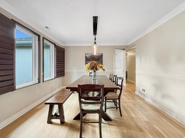 Light wood-finish flooring, two large windows with dark wood plantation shutters, crown molding, and a linear pendant light fixture