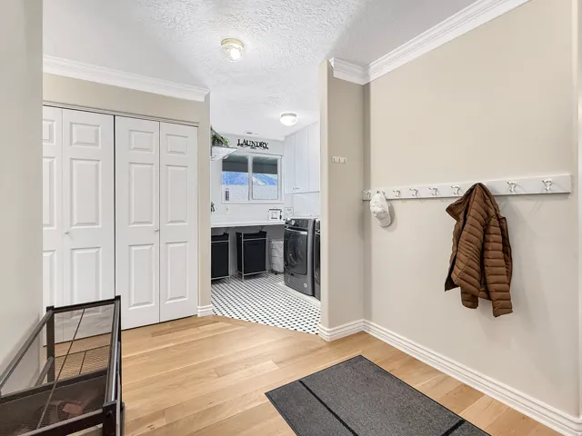 Light wood-finish flooring, white bi-fold doors, crown molding, white wall-mounted coat rack, and black and white patterned flooring in the laundry area