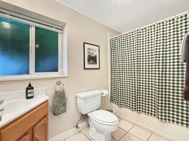 Full bathroom featuring wood-finish vanity with integrated sink, ceramic tile flooring, white porcelain toilet, and checkered shower curtain enclosure