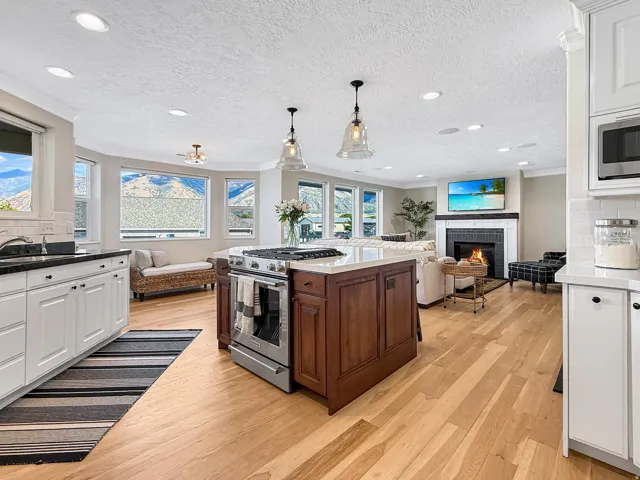 Expansive kitchen and living area featuring wood-finish flooring, a central island with a built-in range, white cabinetry with dark countertops, and a fireplace with a dark surround and white mantle
