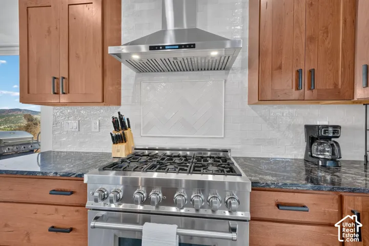 Stainless steel range with a professional-grade hood, wood-finish cabinetry, dark stone countertops, and a white tiled backsplash