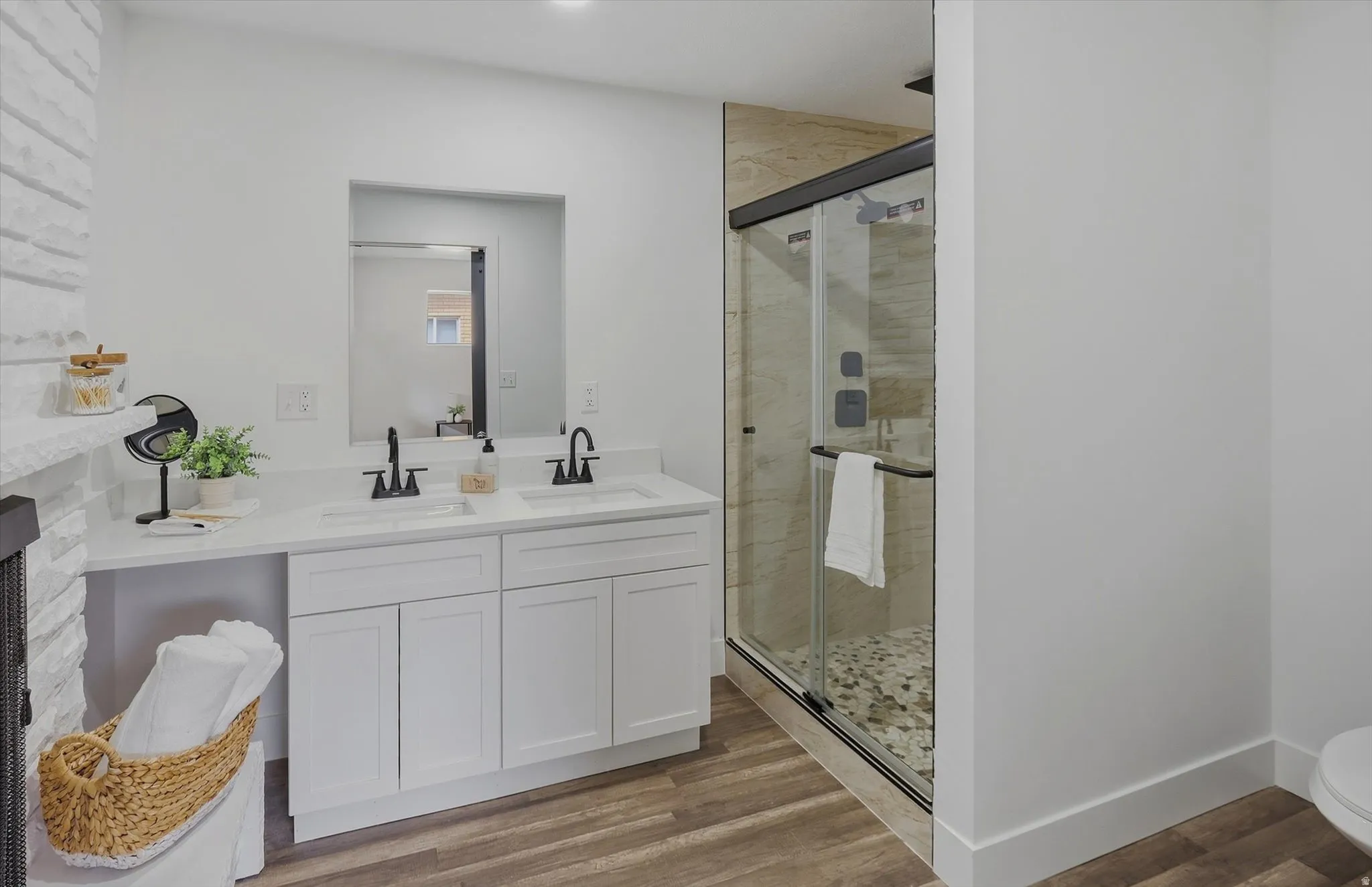 Dual vanity with white cabinetry and integrated sinks, matte black faucets, wood-finish flooring, and a glass-enclosed shower featuring tile walls and a pebble-stone base