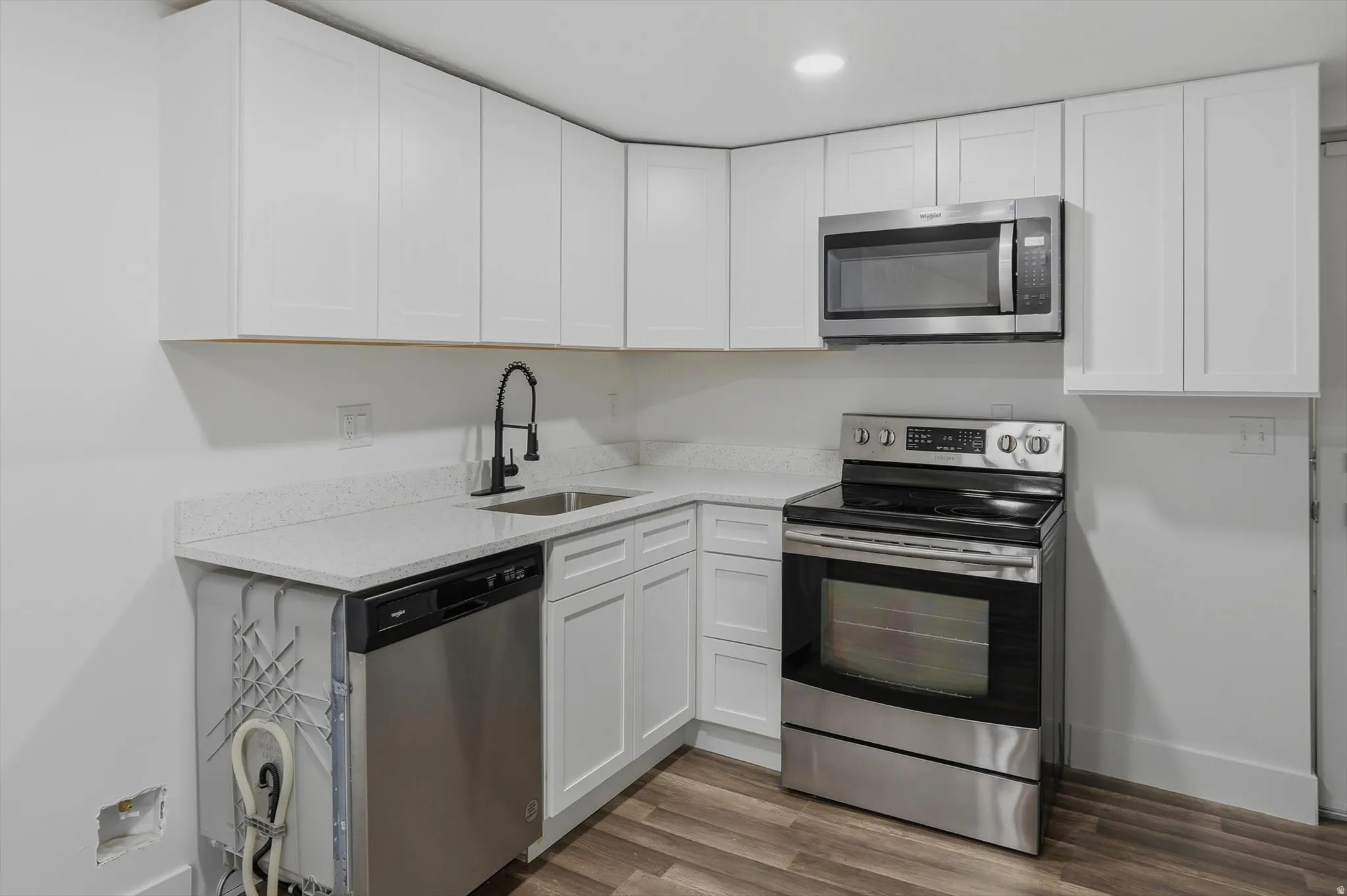 White shaker cabinetry, light-toned countertops, stainless steel appliances, wood-finish flooring, and a matte black gooseneck faucet