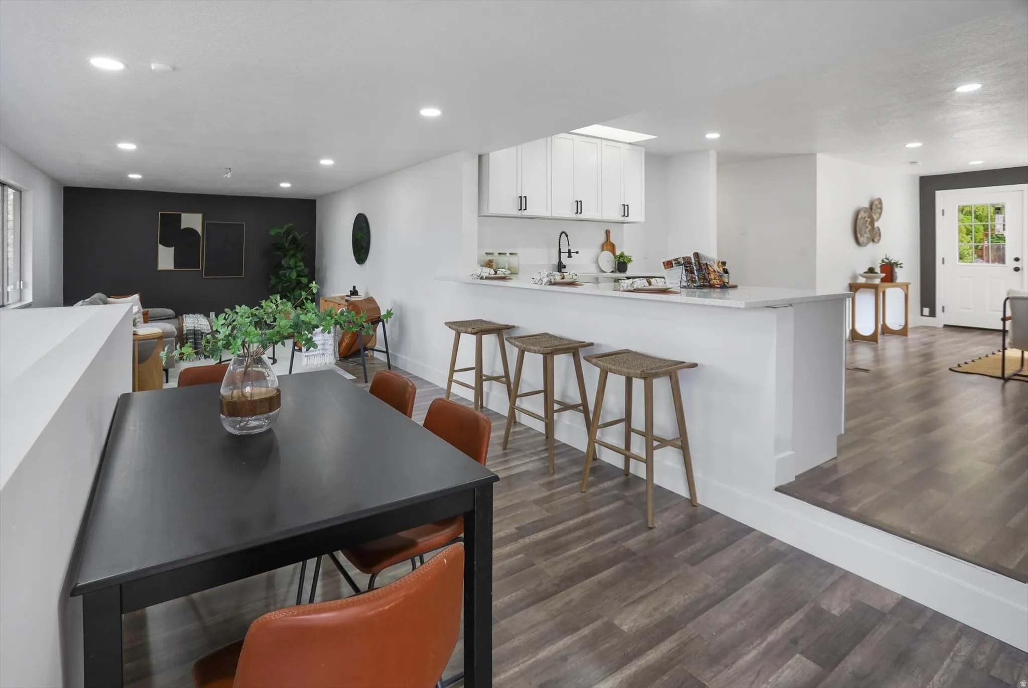 Wood-finish flooring throughout, white kitchen cabinetry with dark hardware, kitchen island with countertop seating, recessed lighting, and a white paneled entry door