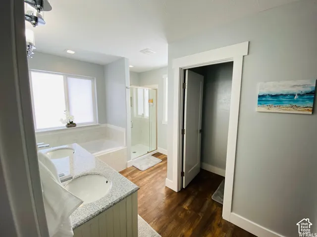 Dual vanity with granite-style countertops, wood-finish flooring, built-in bathtub, glass-enclosed shower, and a large window with privacy coverings