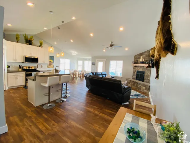 Open-concept living space featuring wood-finish flooring, a kitchen island with seating, a vaulted ceiling with recessed lighting, and a stone fireplace