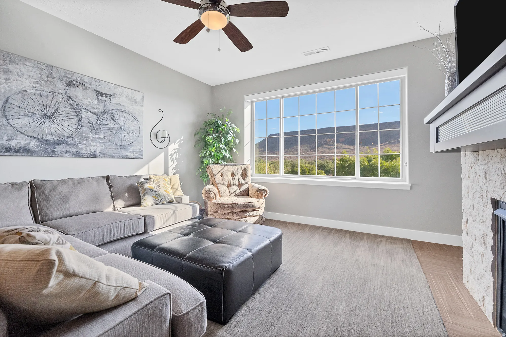 Large multi-pane window with mountain backdrop, ceiling fan with light fixture, fireplace with stone surround and wood mantel, wood-finish flooring