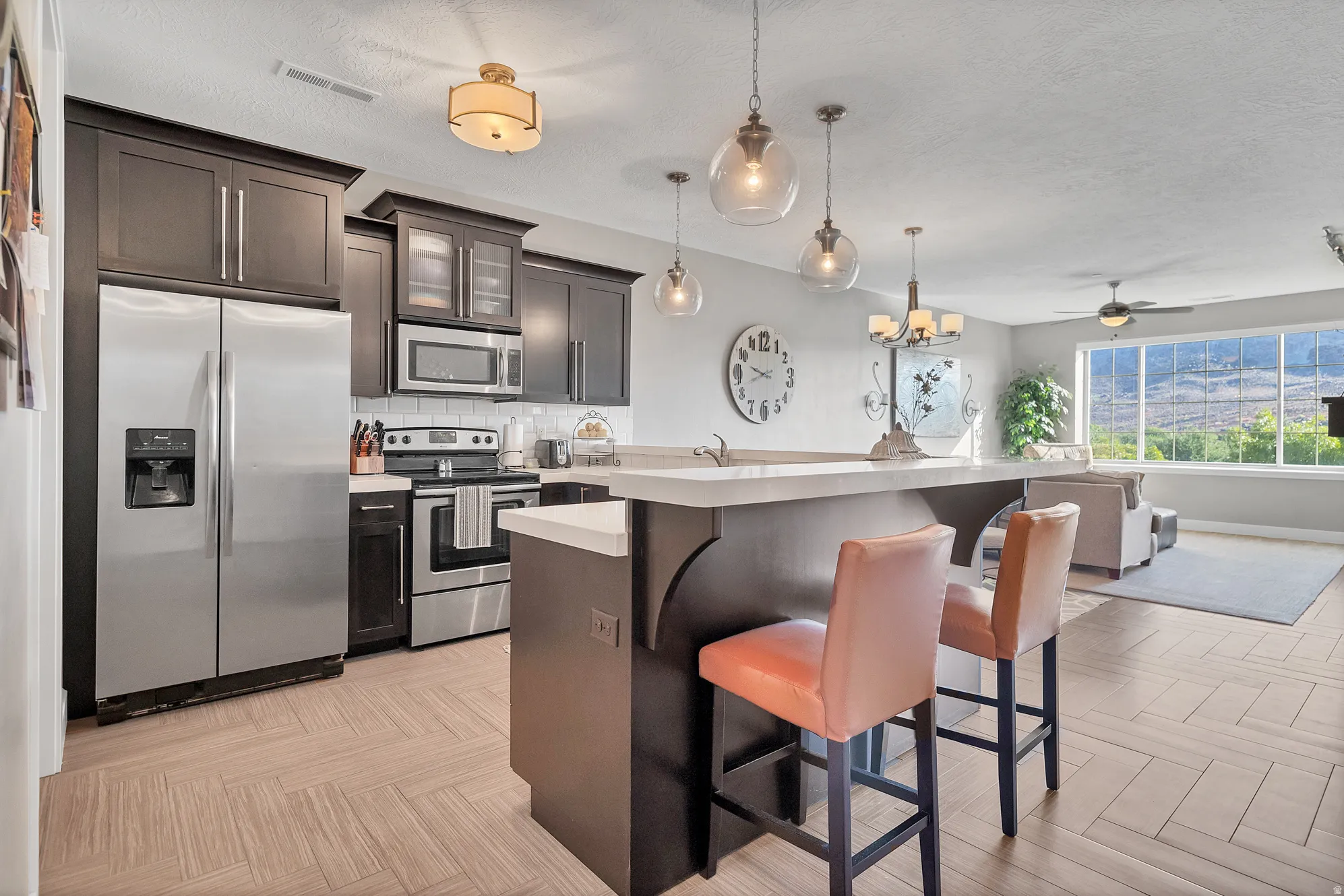 Stainless steel appliances, dark wood-finish cabinetry, light herringbone-patterned flooring, kitchen island with seating, and multiple pendant lights