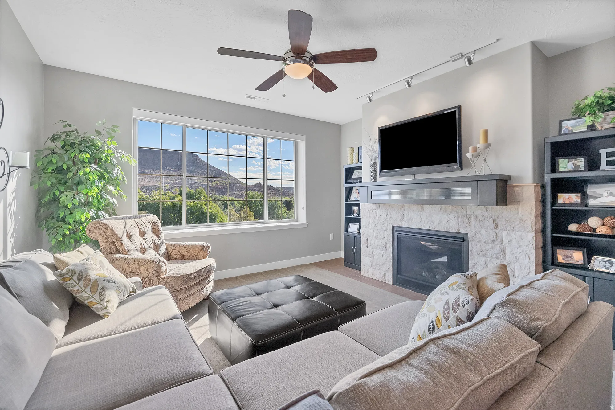 Spacious living area featuring a large picture window, stone-clad fireplace with mantel, ceiling fan, and track lighting
