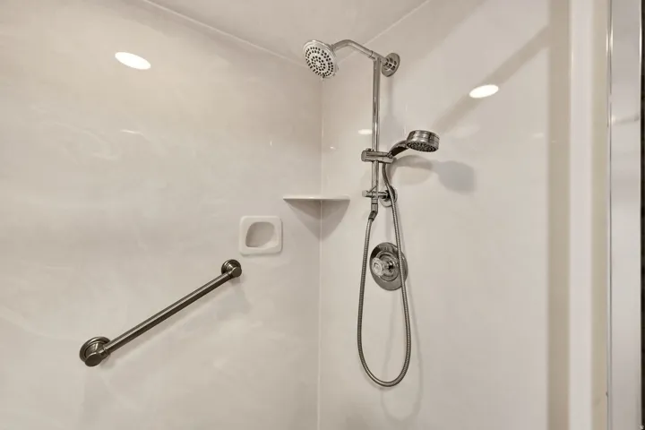 Shower enclosure with white marble-patterned paneling, a rain shower head, a handheld shower fixture, a built-in shelf, and a grab bar