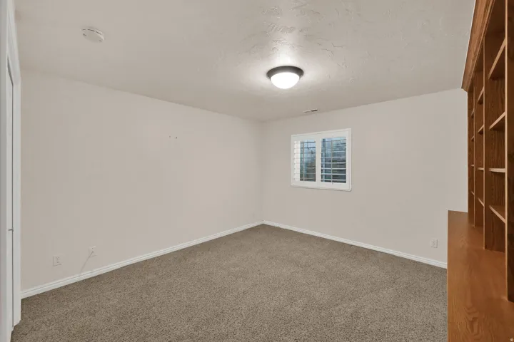 Carpeted room featuring a single window with white plantation shutters, built-in wooden shelving, and an overhead flush-mount light fixture