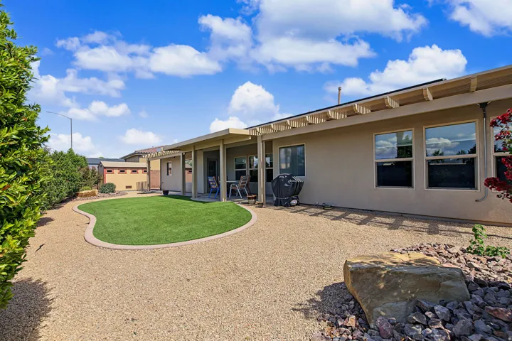 Expansive backyard featuring artificial turf, gravel hardscaping, mature perimeter shrubs, and a covered patio with an exposed beam pergola