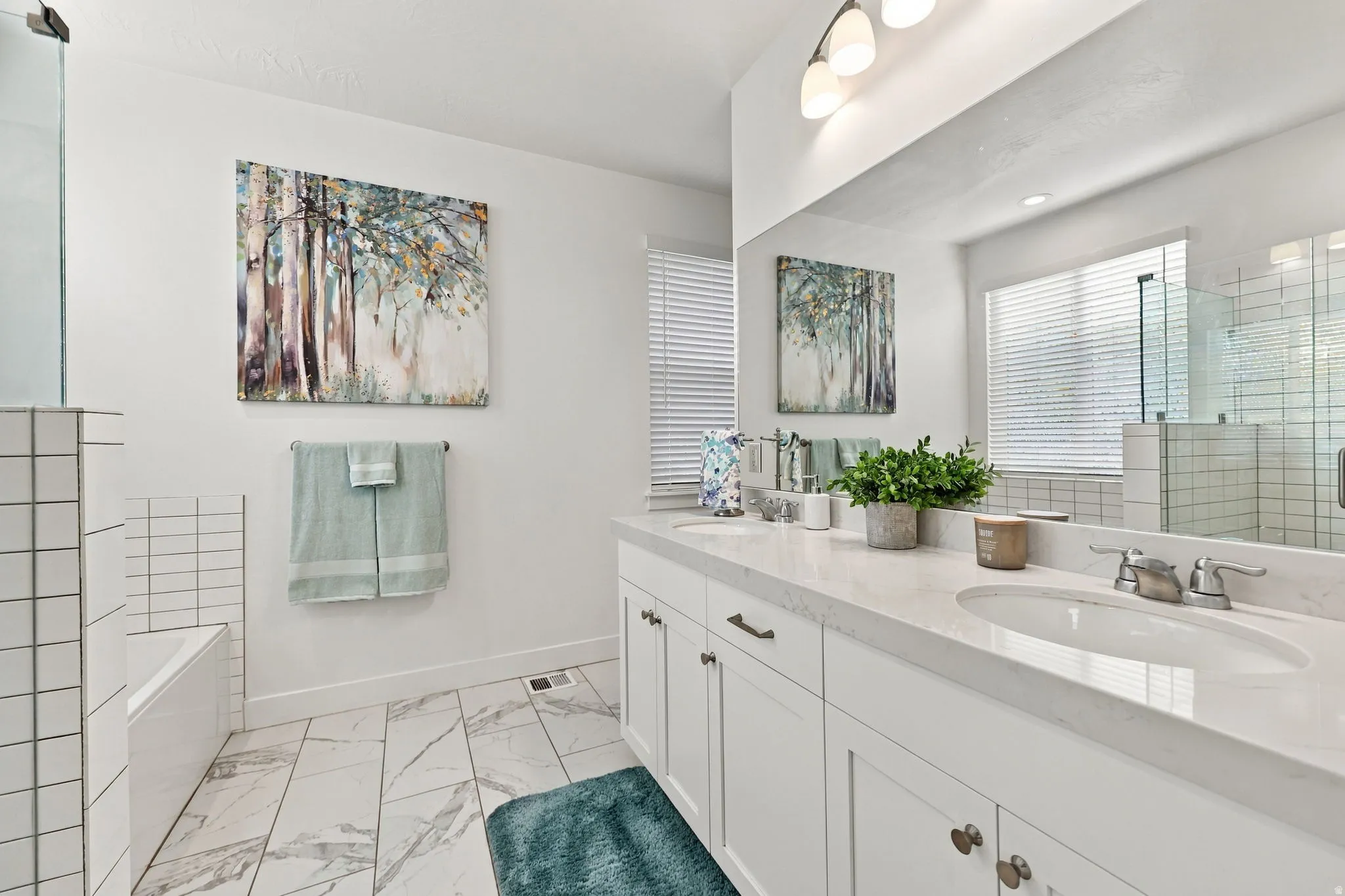 Dual undermount sinks with brushed nickel fixtures, expansive vanity with white shaker cabinetry, large wall-mounted mirror, marble-look tile flooring, and a built-in tub with white subway tile surround