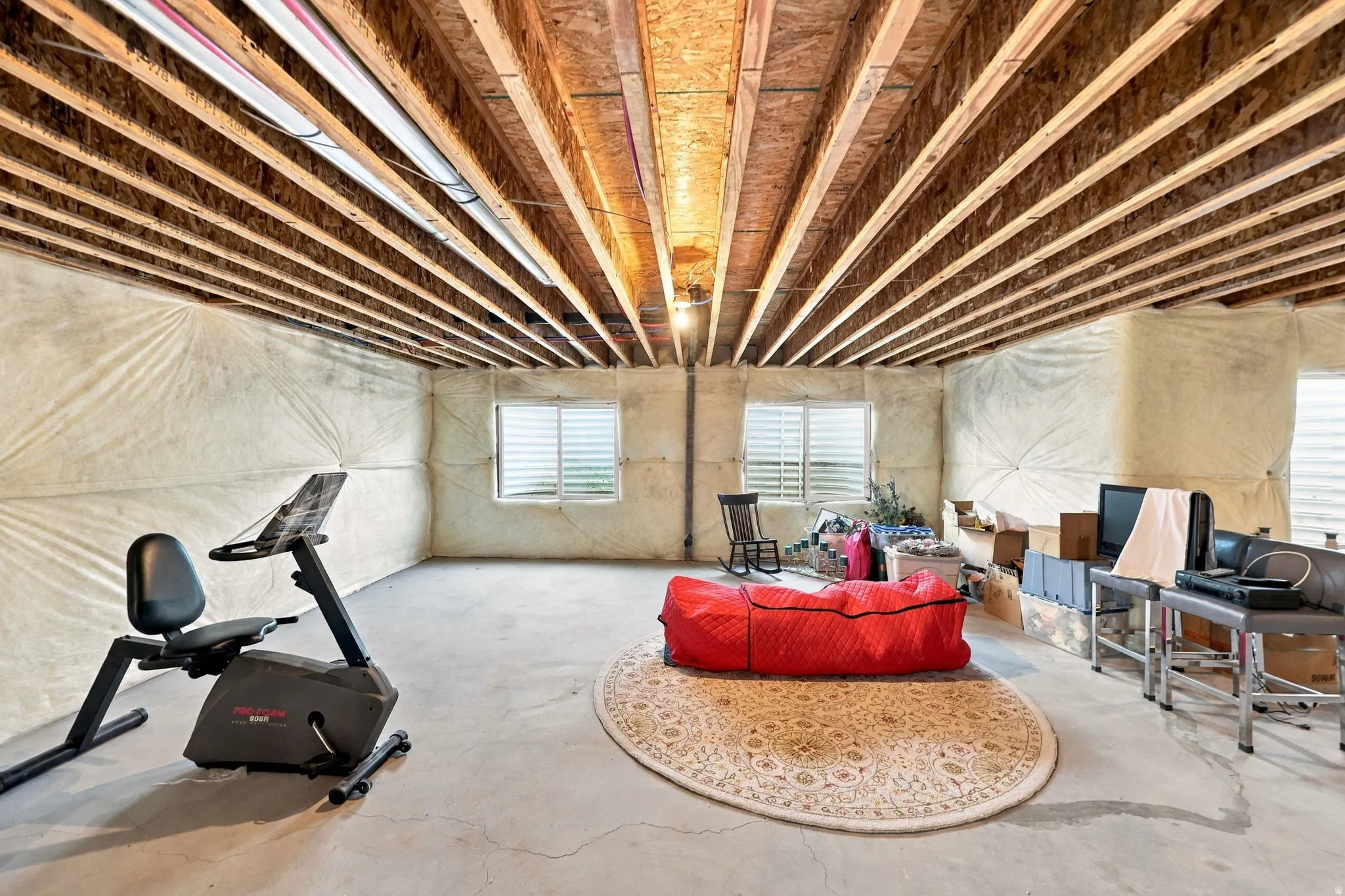 Unfinished basement with exposed wood joists, insulated walls, and two windows with horizontal blinds