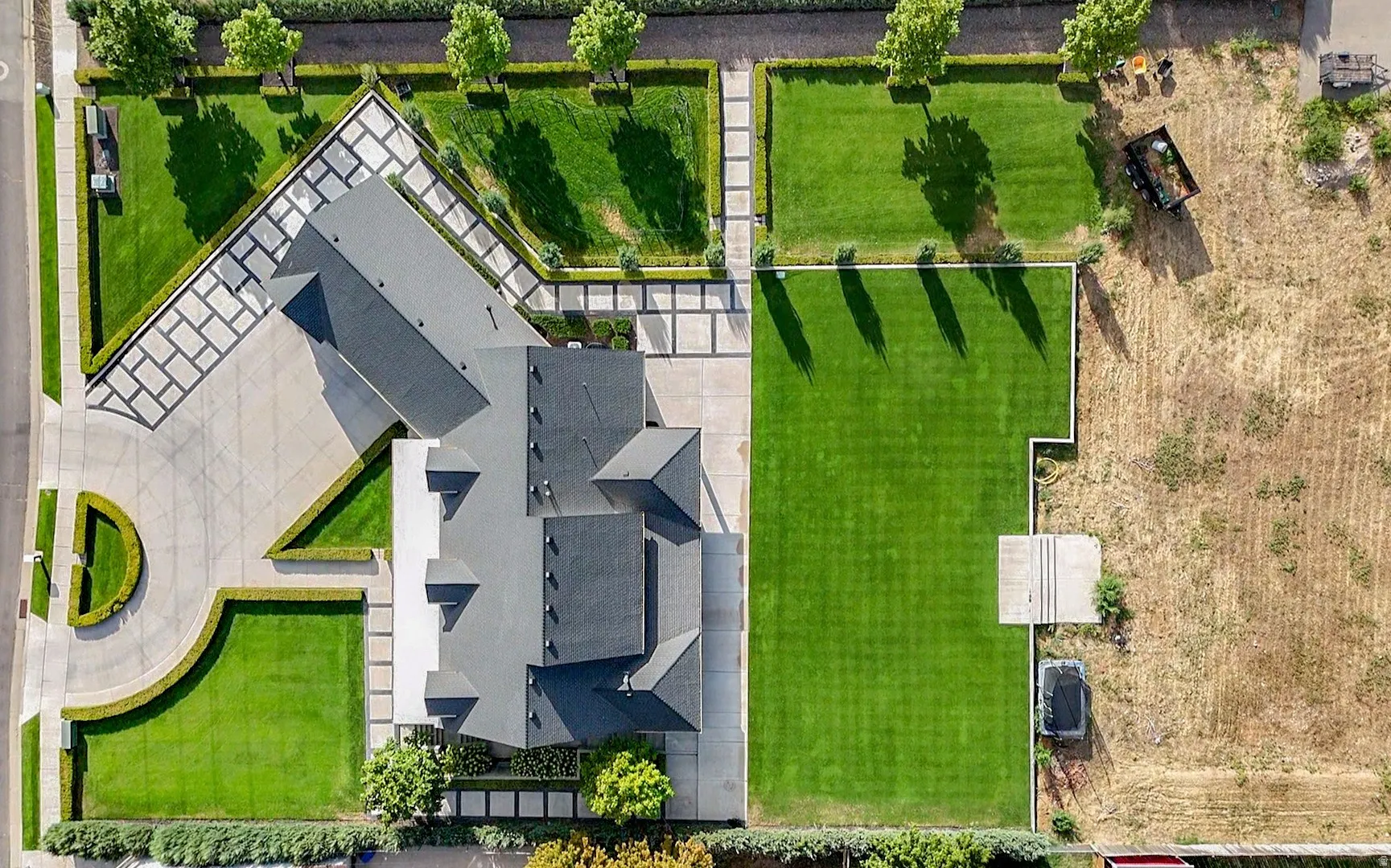 Expansive property featuring a large residence with a dark roof, extensive paved driveways, a circular driveway entrance, manicured lawns, and mature landscaping