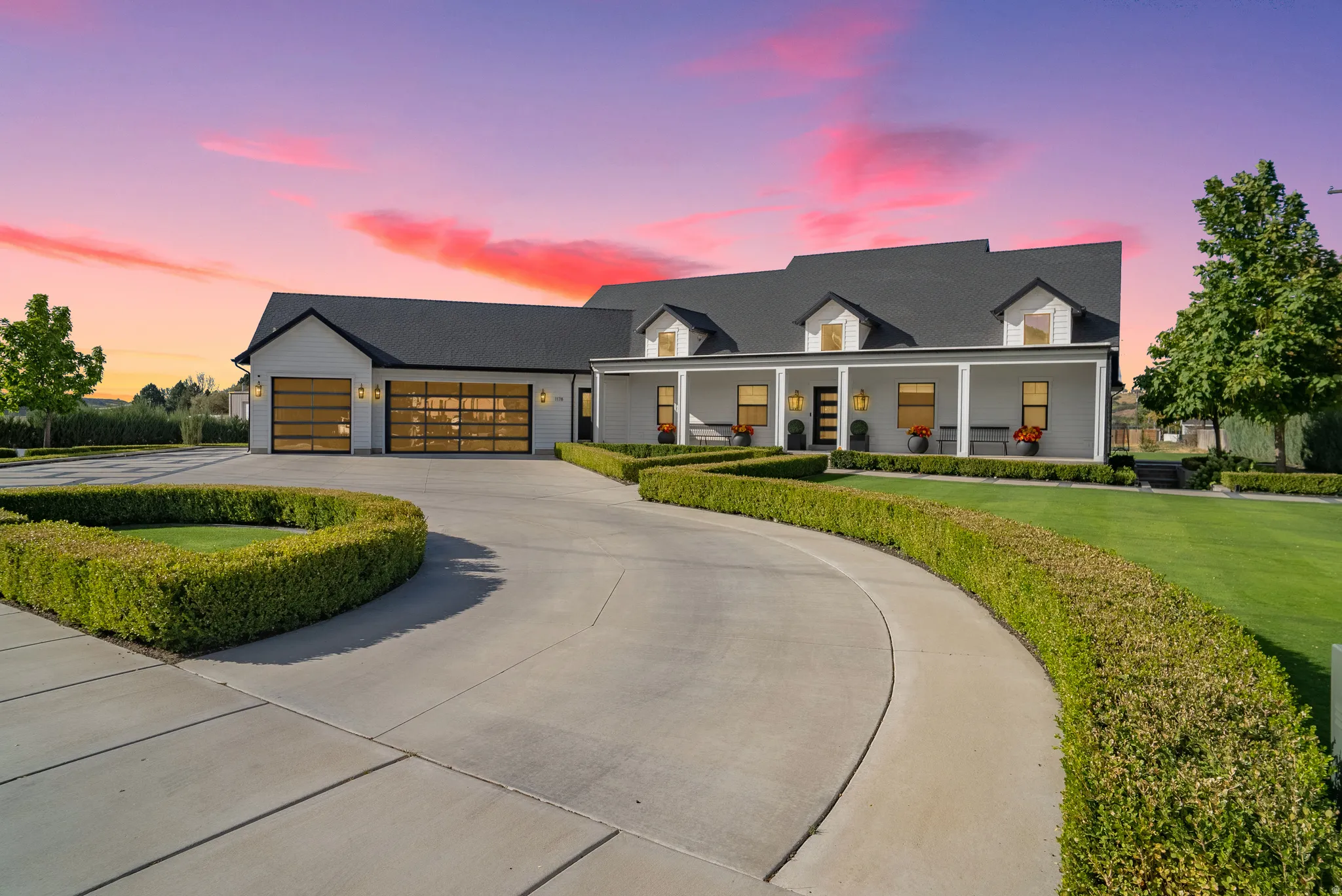 White exterior with black roof, covered front porch, dormer windows, and a circular driveway with manicured hedges