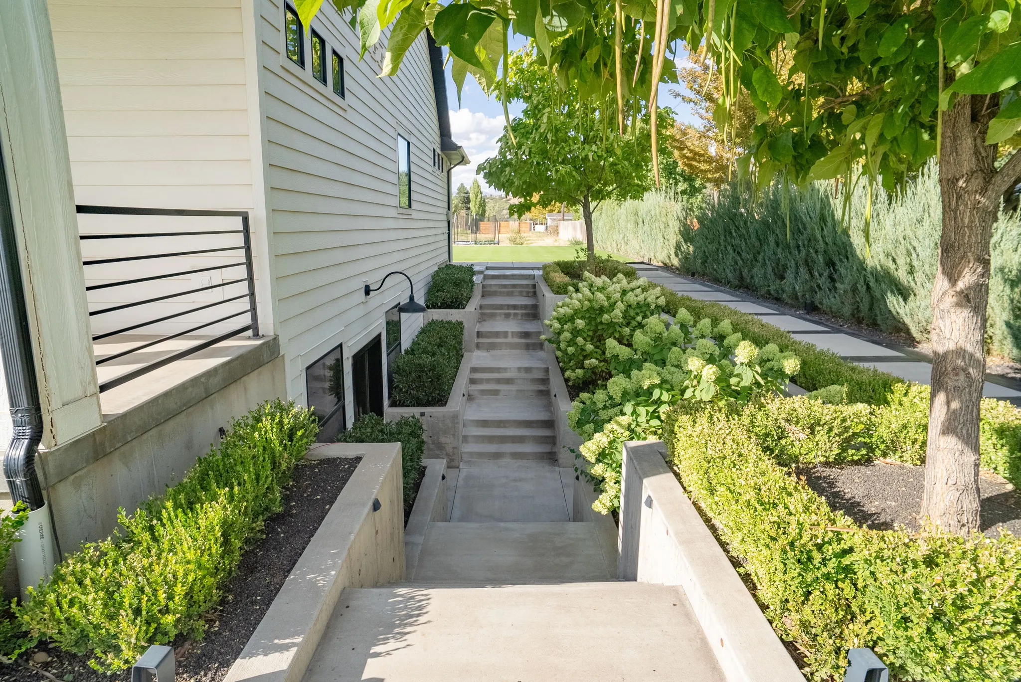 Exterior featuring white horizontal siding, black horizontal railing, concrete steps, built-in planters with shrubs, and mature trees