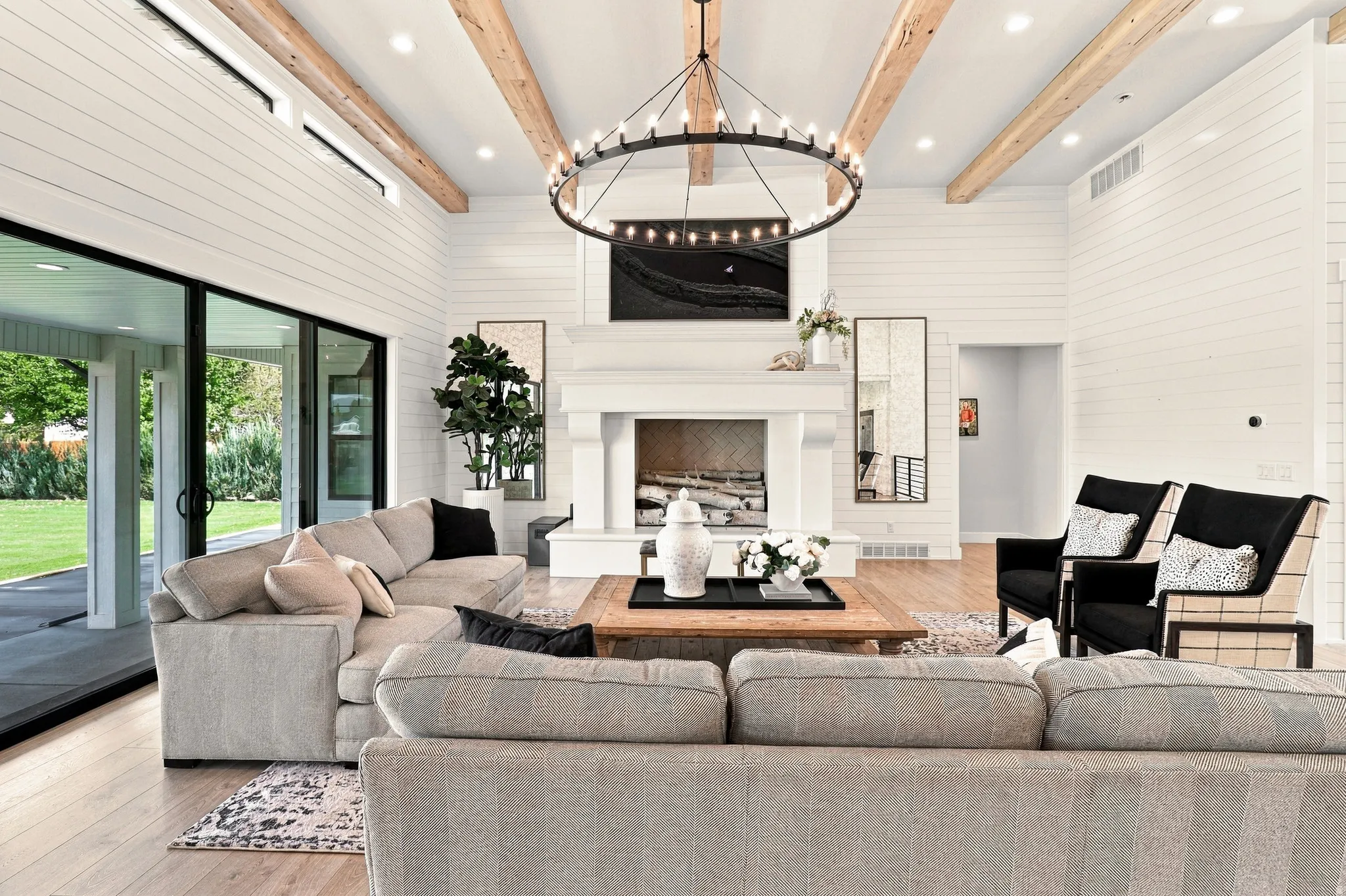Vaulted ceiling with exposed wood beams, white shiplap walls, wood-finish flooring, white fireplace, and black-framed sliding glass doors