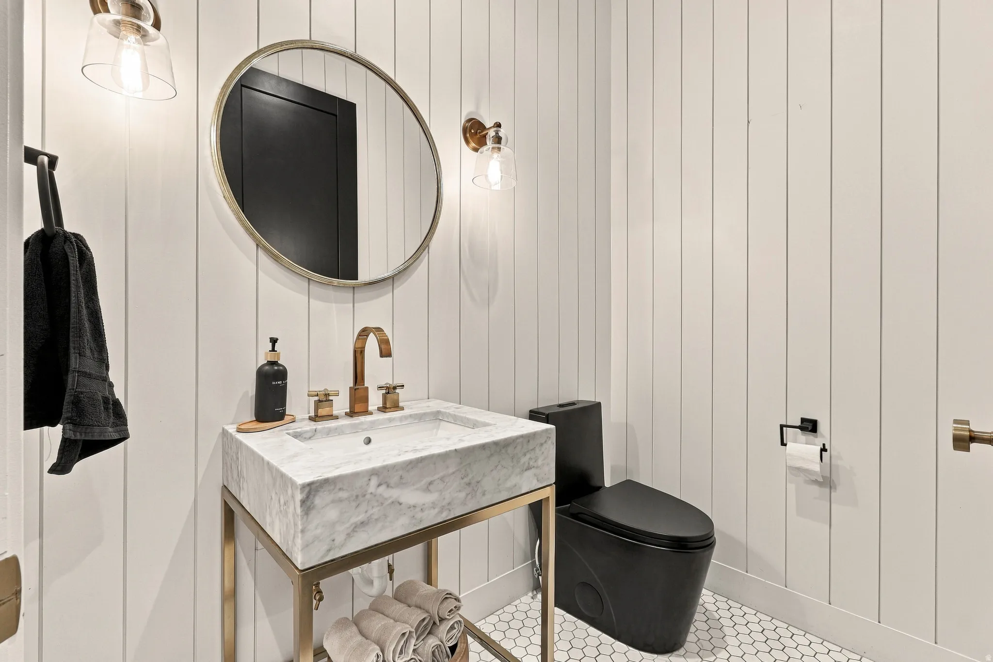 White vertical paneling, hexagonal floor tiling, marble vanity with bronze fixtures, black toilet, and a circular mirror