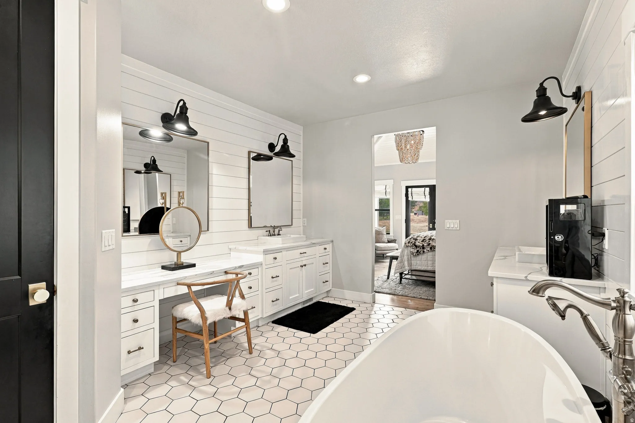 White hexagon tile flooring, shiplap accent wall, dual vanity with undermount sinks, freestanding soaking tub, and black wall-mounted light fixtures