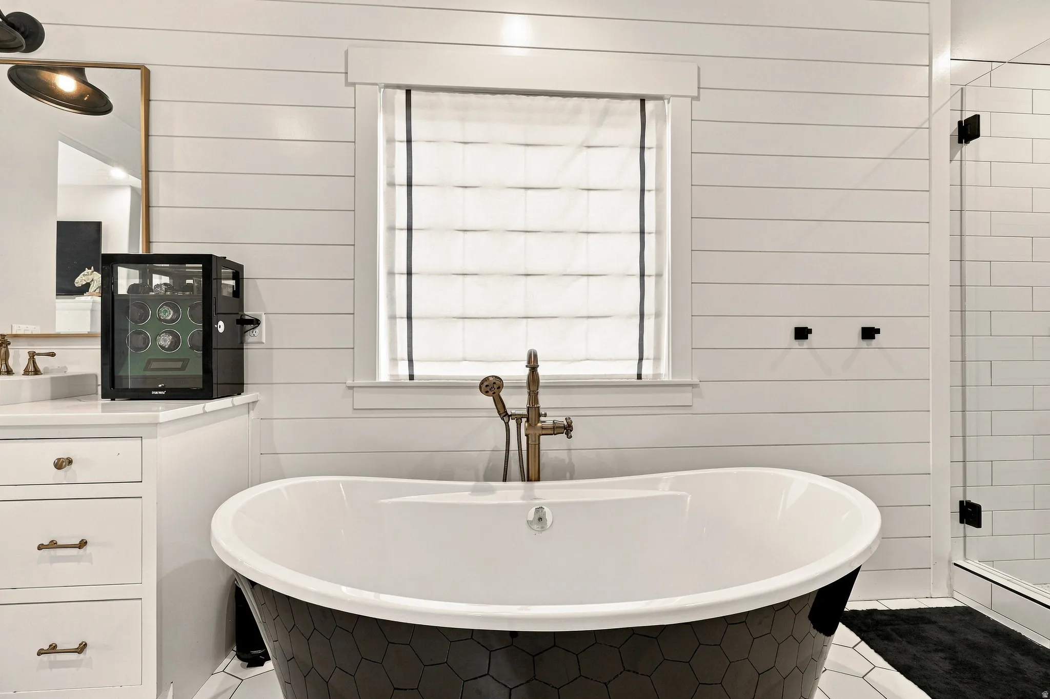 Freestanding soaking tub with a hexagon tile exterior, shiplap wall finishes, frameless glass shower enclosure, and a window with a Roman shade
