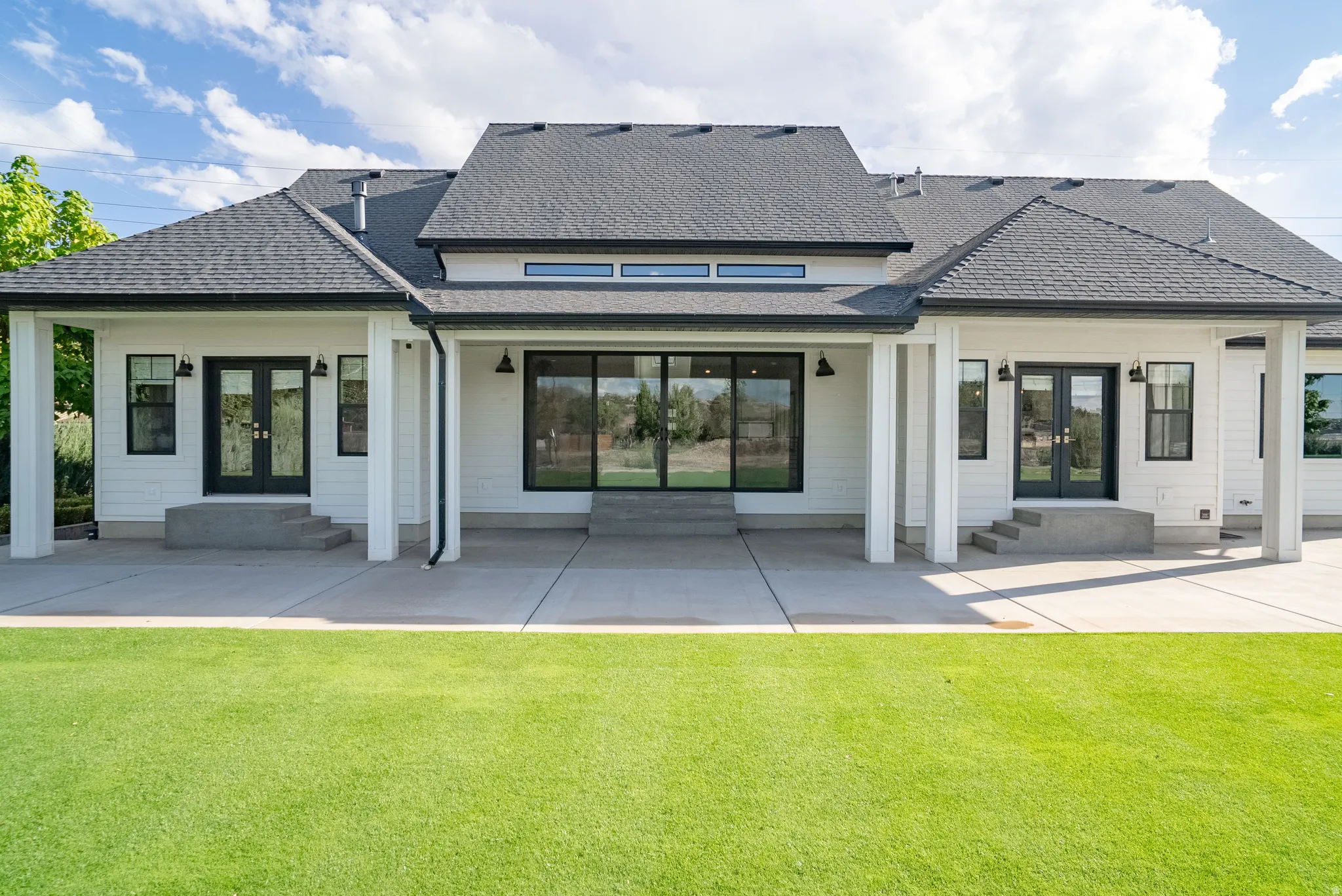 White exterior siding, dark roof, expansive concrete patio, multiple black-framed glass doors, and recessed lighting