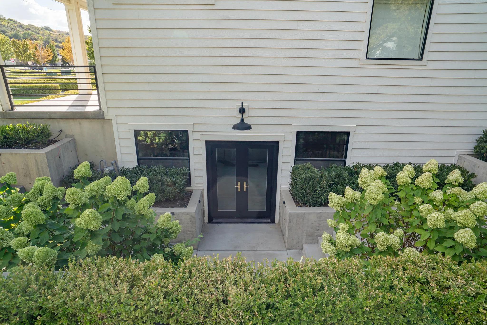White horizontal siding facade with a black double entry door, two ground-level windows, concrete retaining walls, and planted shrubbery