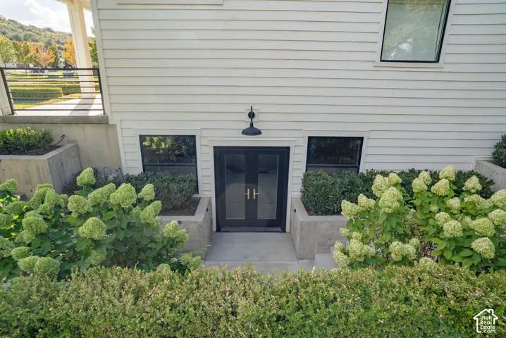 White horizontal siding facade with a black double entry door, two ground-level windows, concrete retaining walls, and planted shrubbery