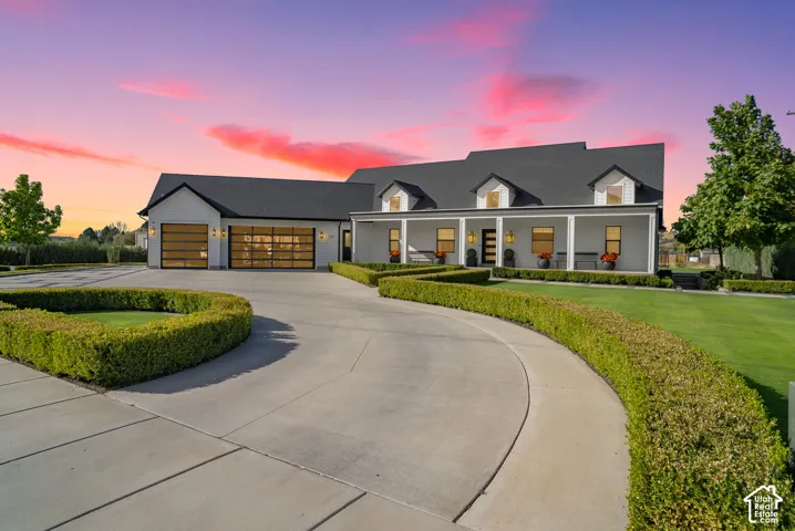 White exterior with black roof, covered front porch, dormer windows, and a circular driveway with manicured hedges