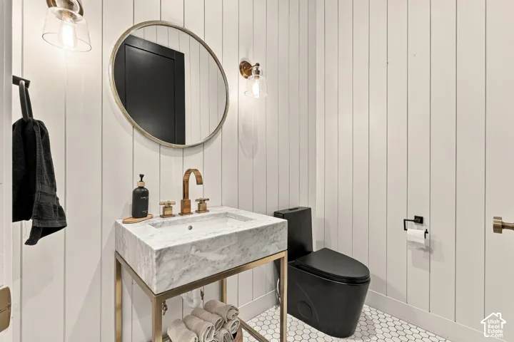 White vertical paneling, hexagonal floor tiling, marble vanity with bronze fixtures, black toilet, and a circular mirror