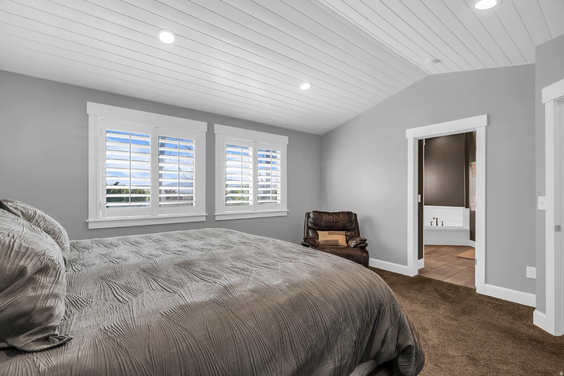 Vaulted shiplap ceiling with recessed lighting, dual windows with plantation shutters, with an adjacent bathroom suite