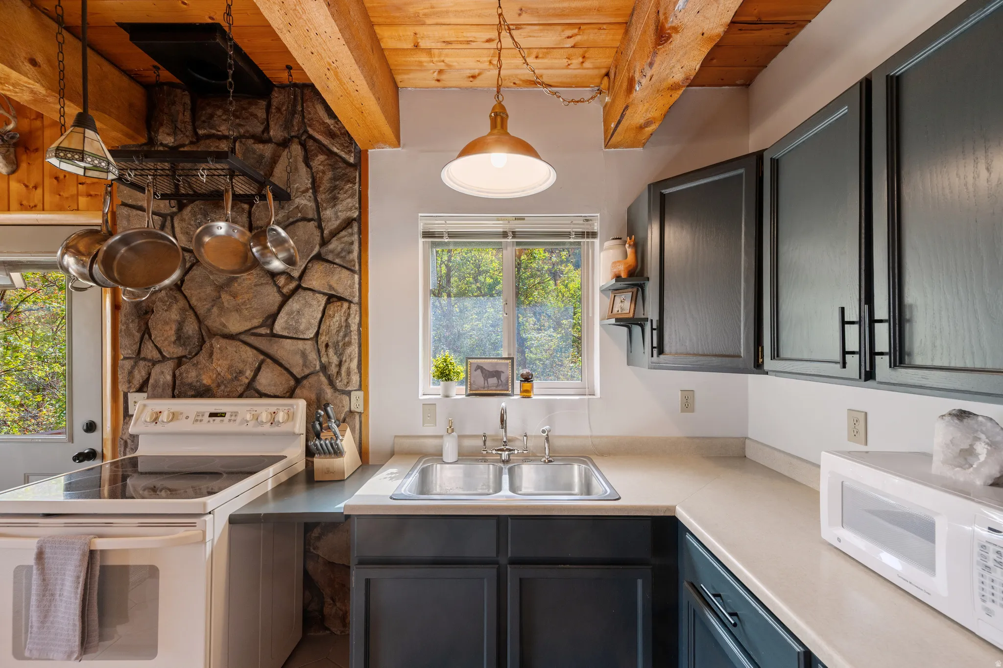 Kitchen featuring a stone accent wall, wood-paneled ceiling with exposed beams, dark cabinetry with bar pulls, a double basin sink, and a smooth-top range