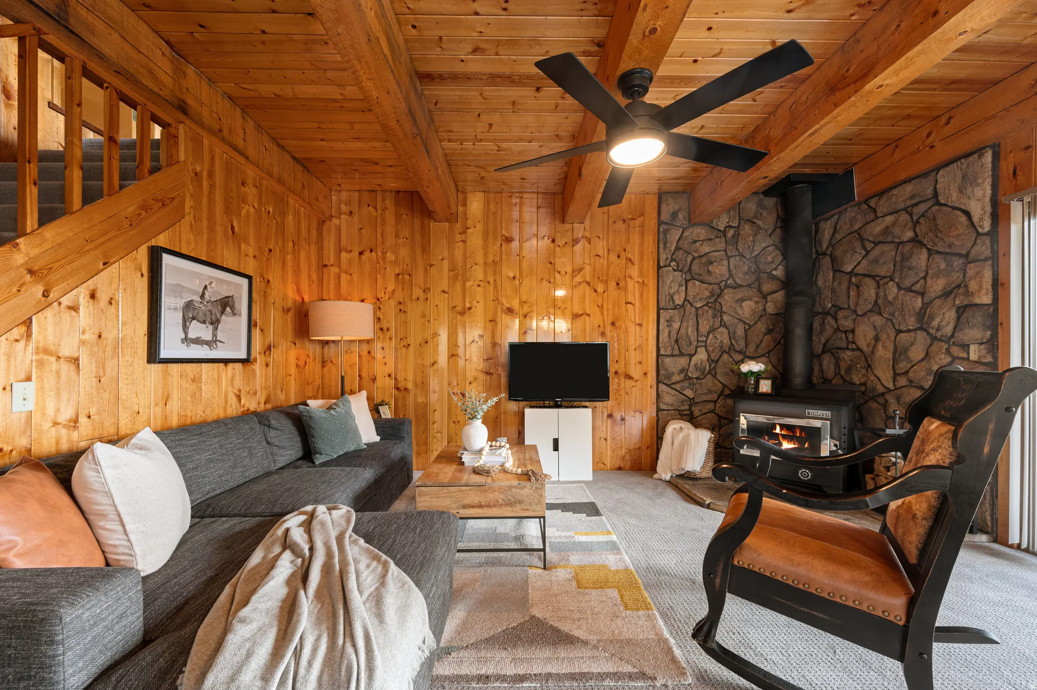 Rustic wood-paneled living area featuring exposed beam ceiling, ceiling fan with integrated lighting, wood-burning stove with stone surround, and comfortable seating arrangement