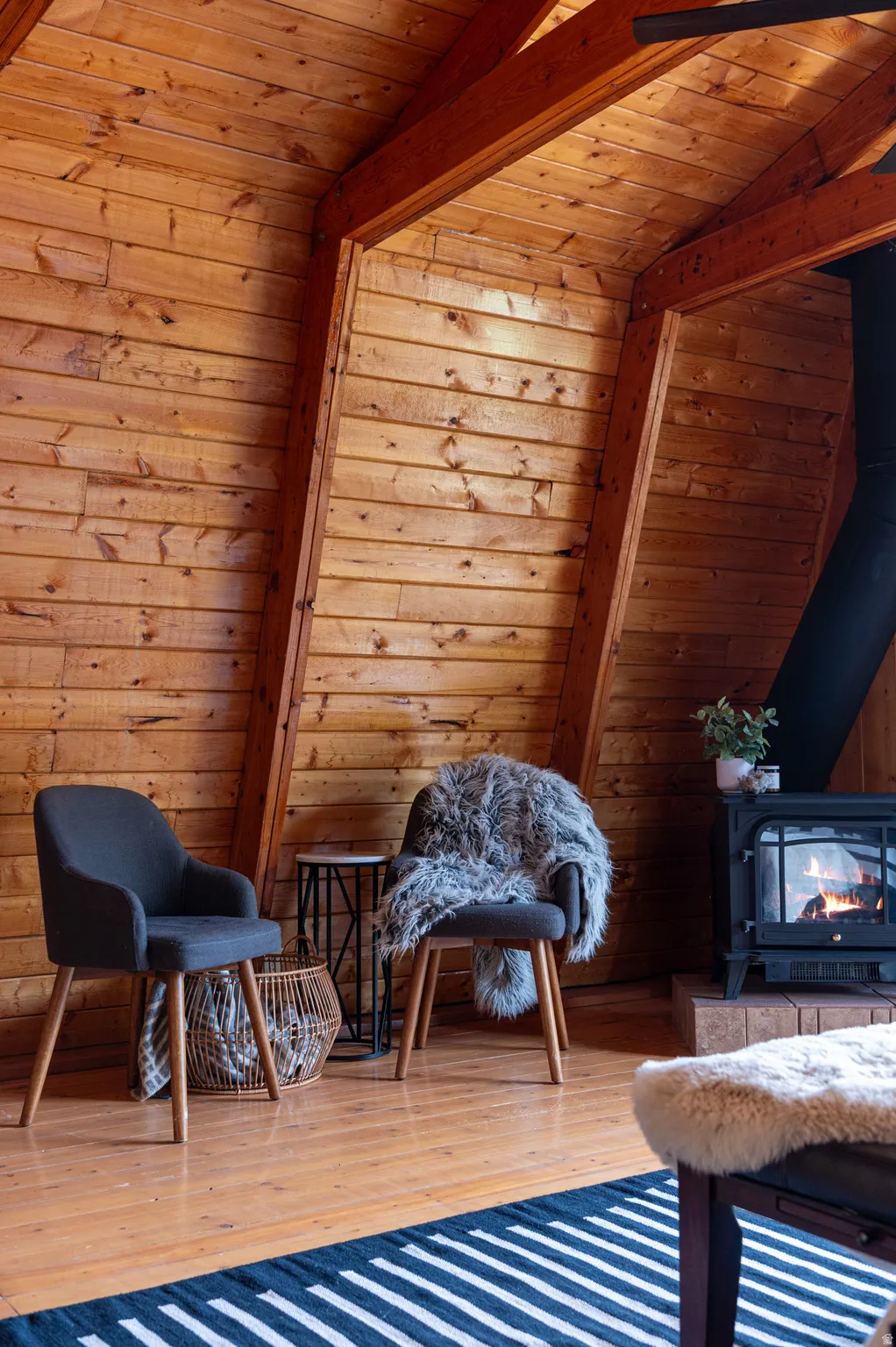 Slanted wood panel walls, exposed wood beams, wood-finish flooring, and a black wood-burning stove