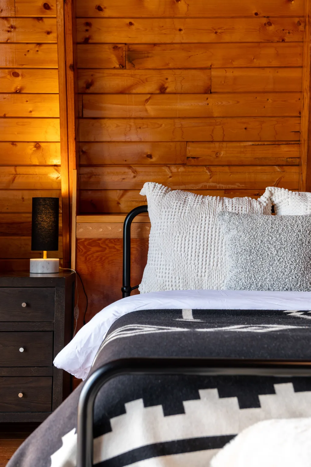 Wood-paneled wall, dark wood-finish three-drawer nightstand, and black metal bed frame