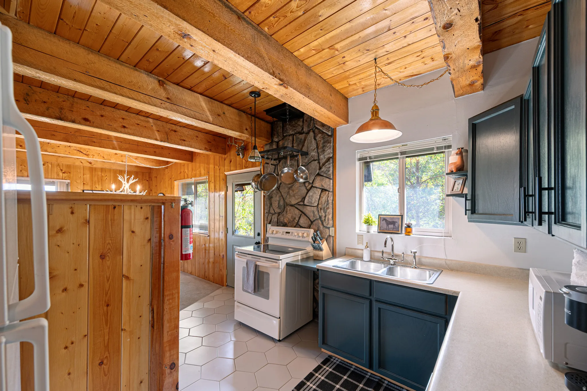 Exposed wood beam ceiling, stone-clad chimney feature, dual basin stainless steel sink, white hexagonal tile flooring, and dark blue cabinetry