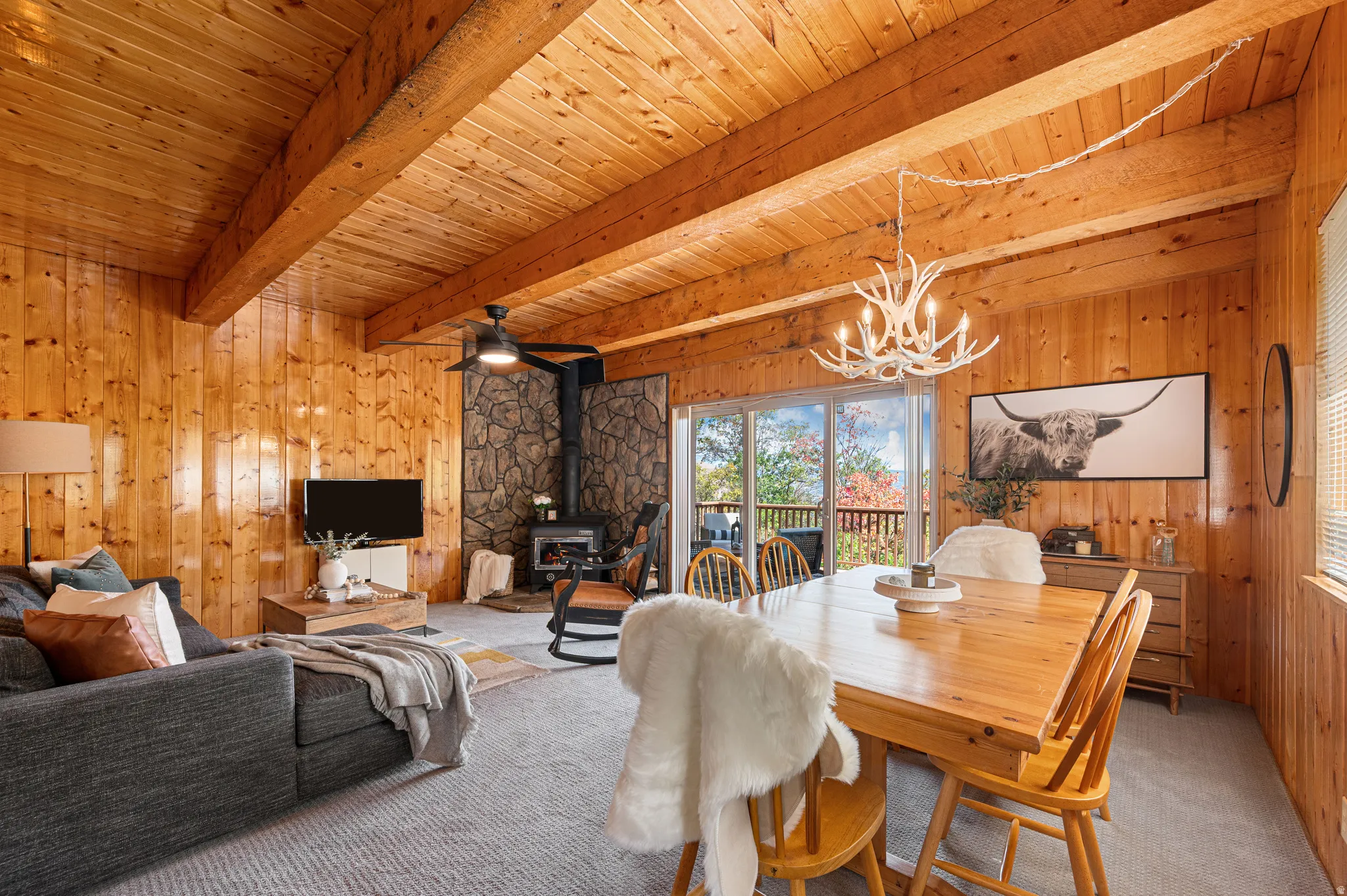 Vaulted wood-paneled ceiling with exposed beams, wood-paneled walls, stone-clad fireplace surround with wood-burning stove, and sliding glass doors to outdoor space