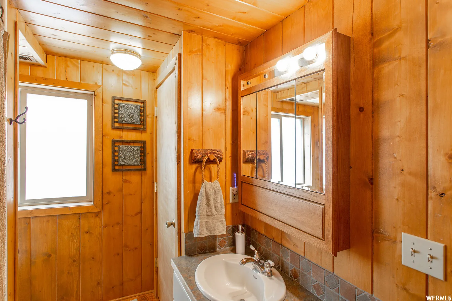 Wood-paneled walls and ceiling, built-in medicine cabinet with integrated lighting, window for natural light, and a vanity with an integrated sink