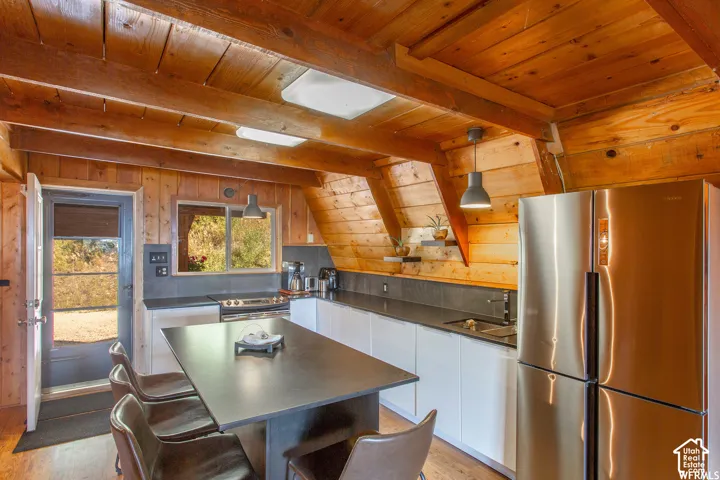 Wood-paneled ceilings with exposed beams, stainless steel refrigerator, white flat-panel cabinetry, dark countertops, and wood-finish flooring