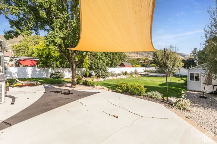 Expansive concrete patio with a shade sail, large green lawn, mature trees, and a white privacy fence