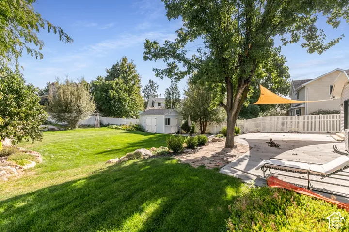 Expansive backyard featuring a manicured lawn, mature trees, white picket fencing, a dedicated storage shed, and a concrete patio