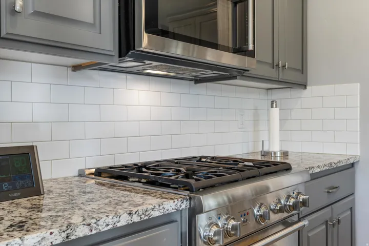 Stainless steel gas range with grates, granite-finish countertops, white subway tile backsplash, gray cabinetry with stainless hardware, and a built-in stainless steel microwave