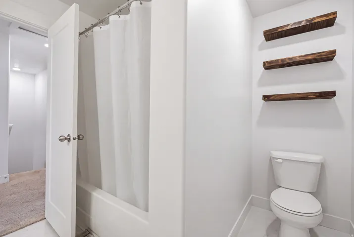 Tub/shower combination with a white curtain, white toilet, and three wall-mounted wood-finish shelves