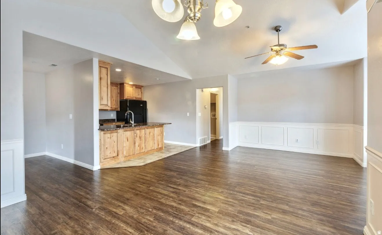Vaulted ceiling, wood-finish flooring, wainscoting, island with wood cabinetry, and a ceiling fan with integrated lighting