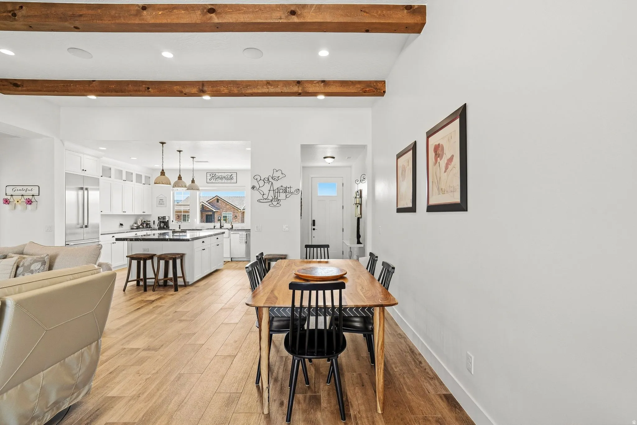 Open-concept living area with exposed wood beams, wood-finish flooring, recessed lighting, and a white kitchen featuring an island with counter seating