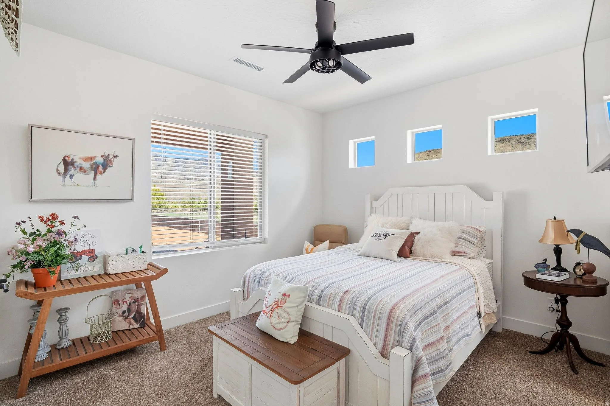 White paneled bed frame, large window with blinds, three upper rectangular windows, black ceiling fan, and carpeted flooring