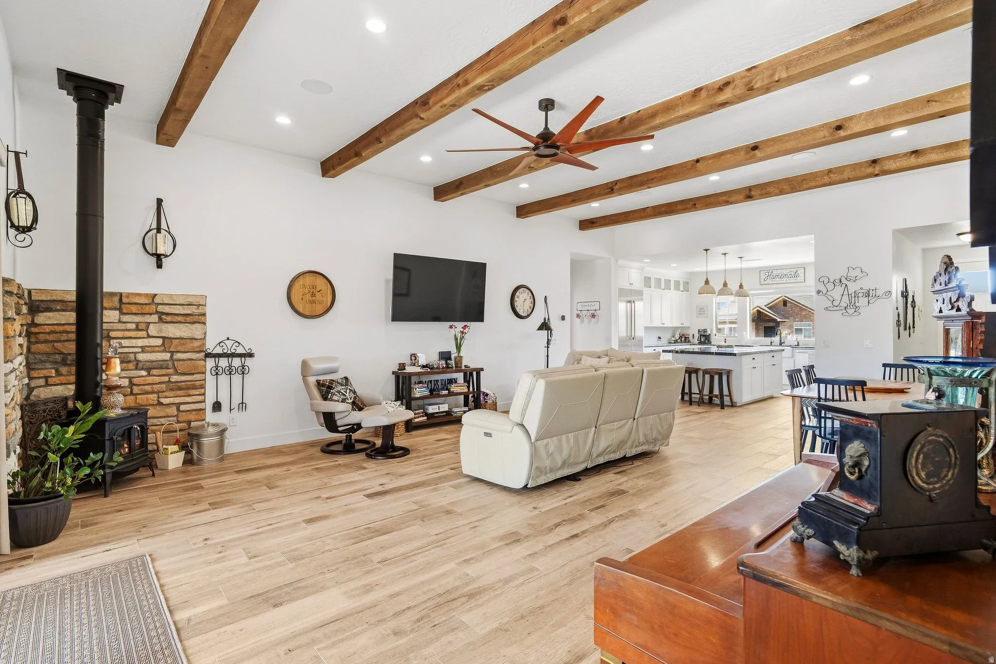 Exposed wood beam ceiling with recessed lighting, wood-finish flooring, stone-clad fireplace with a black wood-burning stove, white kitchen cabinetry with an island, and pendant lighting