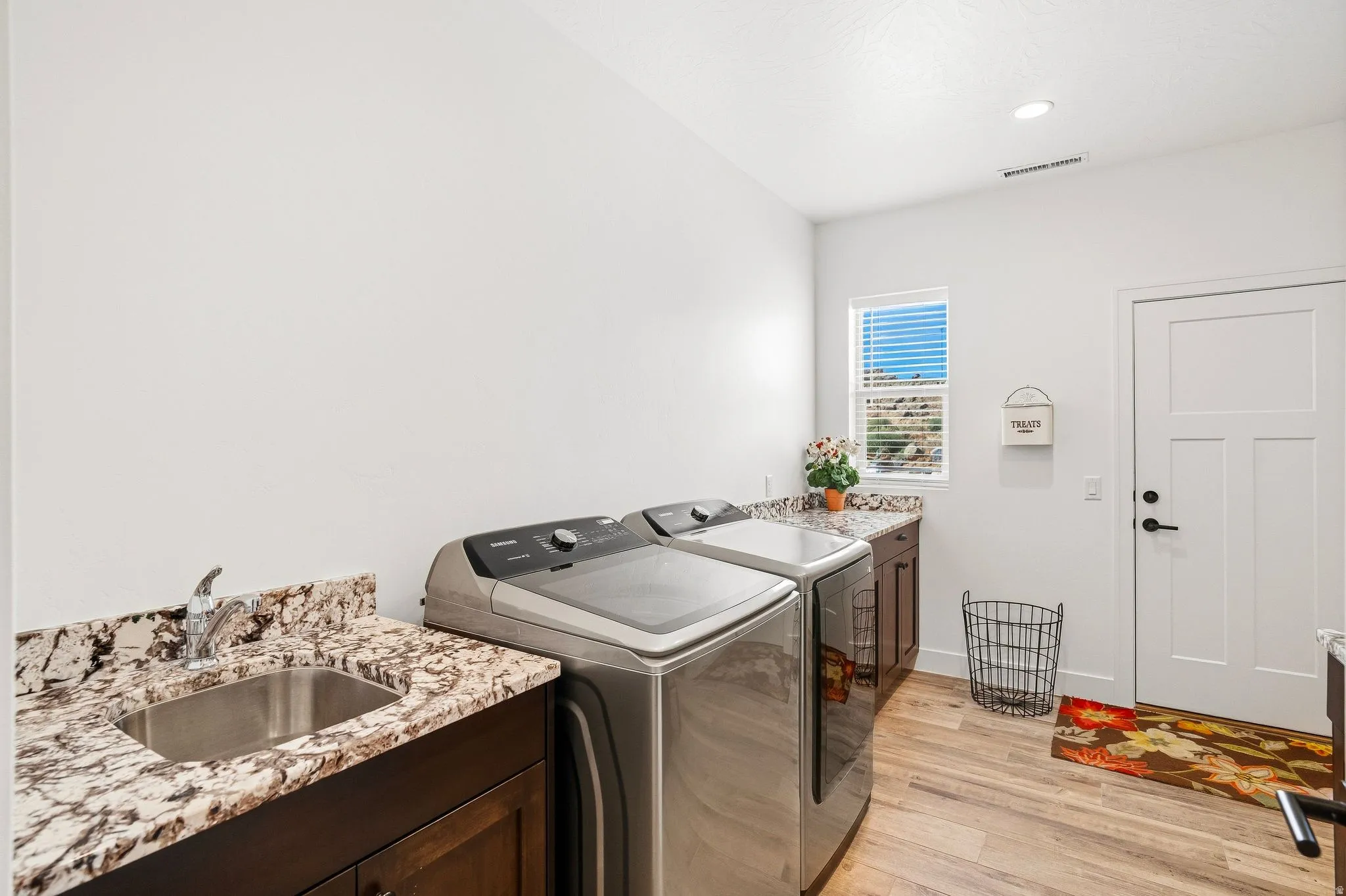 Laundry area featuring wood-finish flooring, a utility sink with a chrome faucet, dark wood cabinetry, and speckled countertops